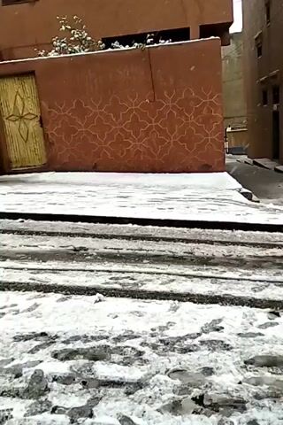 The video shows a street covered in a thin layer of snow. The buildings lining the street are primarily multi-story and painted in shades of tan and orange. The snow is patchy, revealing the dark asphalt of the street beneath. Tire tracks are visible in the snow. Several people are walking on the street, including one person in a dark jacket and another pulling a sled. A car is parked on the street, partially covered in snow. The sky is overcast.