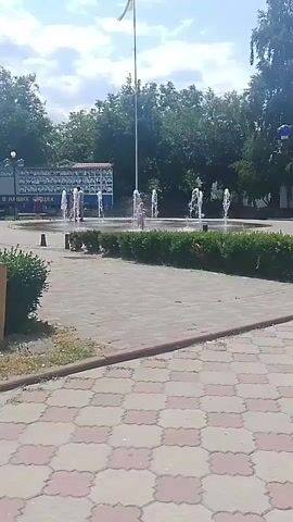 A video shows a park with a fountain and trees. A flagpole with a flag is visible in the background. There are people in the fountain. The ground is paved with hexagonal tiles. The weather appears to be sunny with some clouds in the sky.