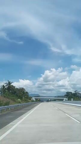 A car is traveling on a highway under a bright blue sky with fluffy white clouds. The road has multiple lanes and is lined with guardrails and vegetation. An overpass crosses the highway in the distance, and other cars are visible on the road. The scene suggests a sunny day suitable for travel.