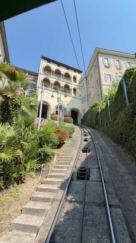 A low-angle, first-person perspective shot shows a funicular railway track ascending a steep incline. The tracks are made of metal, and a concrete path runs alongside them. Several large black wheels are visible attached to the trackbed. To the left of the tracks, a set of concrete stairs climbs upwards, bordered by lush green bushes and palm trees. To the right and center, two buildings stand on either side of the incline. The building on the left is taller and features multiple arched balconies. The building on the right is lighter in color and has visible windows. Overhead, power lines for the funicular are stretched across the bright blue sky. The overall environment appears to be a sunny, outdoor location, likely a tourist destination. No people are directly visible in the frame.