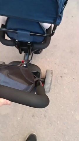 The video shows a close-up, first-person perspective of someone pushing a blue and black tricycle with a canopy. The tricycle is on a paved surface, possibly a sidewalk or road, with some debris visible. A person's hand, wearing a white sleeve, is seen holding the handle of the tricycle. The person's feet, wearing black and white sneakers, are also visible at the bottom of the frame. The scene appears to be outdoors, and based on the lighting, it is likely daytime. The overall mood of the video is neutral and observational. No text is visible in the video.