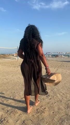 A woman is walking barefoot on a beach. She is wearing a sheer black dress and carrying a straw hat in her right hand. She has long, dark, curly hair. Behind her is a volleyball net and some beach chairs and umbrellas. The sky is blue and the sand is light colored.