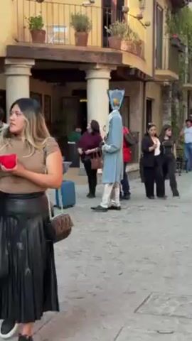 The video shows a person dressed in a light blue costume, standing on a cobblestone street. The costume includes a unique headpiece resembling a stylized lantern or tower. The person is surrounded by other people, some of whom are taking pictures with their phones. In the background, there is a building with columns and balconies. A blue suitcase sits on the street near a table. The scene appears to be in a public space, possibly a town square or outdoor area of a restaurant.