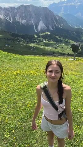 A young woman stands in a meadow filled with yellow flowers, with a mountain range in the background. She is wearing a pink tank top and grey shorts. A person's hand extends into the frame, offering her a single yellow flower. She smiles and brings her hands to her mouth in a gesture of surprise and delight. The video captures a romantic moment set against a beautiful natural landscape.