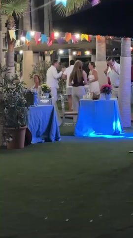 The video captures an outdoor evening event, likely a party or celebration, set against a backdrop of palm trees and a string of colorful flags. The ground is covered in artificial turf. In the foreground, two round tables are draped with light blue tablecloths. The table on the left has a potted plant and various items on it, while the table on the right has more items, including flowers and what appears to be a drink. Blue uplighting illuminates the scene.

Several people are visible. In the center, a man in a white shirt and shorts is interacting with a woman wearing a white top and cream-colored pants. To the left, a man in a white shirt and pants is speaking with a woman sitting at a table. On the right, a man in a white shirt and shorts is standing and gesturing. Further back, other guests are present, some of whom are standing or sitting. The overall atmosphere appears festive and social, with people engaged in conversation and enjoying the evening.