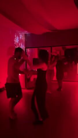 The video shows two people dancing in a room lit with red light. The man is wearing a short-sleeved shirt and shorts, while the woman wears a black dress with a slit. They are holding hands and moving to the music. Other people can be seen in the background, some standing, and one couple also dancing. In the background, there's a DJ booth, a mirror, and some decorations on the wall.
