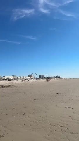 The video captures a wide shot of a sandy beach on a clear, sunny day. In the background, a line of white buildings stretches across the horizon, with a Ferris wheel visible to the right. The beach itself is mostly empty, with a few scattered items of debris and some people in the distance. The foreground of the video is dominated by the textured sand, showing ripples and footprints.

At the 00:01 mark, a shirtless man wearing olive green cargo pants and a white baseball cap walks towards the camera, his back to the viewer. He is barefoot.

At the 00:02 mark, a small child wearing a white shirt and blue shorts is seen walking away from the camera, further down the beach from the man.

The video continues with shots of the beach and the buildings in the background, with no further significant action or interactions observed.
