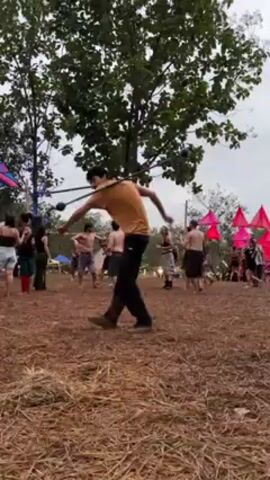 A man in a yellow shirt and dark pants holds a long stick and performs a dance or rhythmic exercise, twirling and moving the stick around his body. He is surrounded by a group of people dancing or moving rhythmically on a ground covered in straw or dry grass.  The scene appears to be at an outdoor gathering or festival, with colorful flags or decorations visible in the background.  The area is somewhat open, with scattered trees and a cloudy sky.