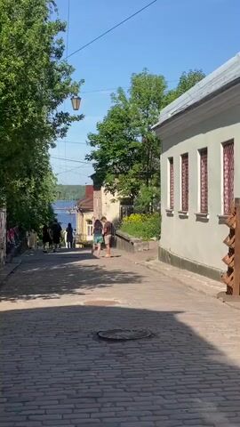 The video shows a cobblestone street with buildings on either side. There are trees lining the left side of the street, and a body of water can be seen in the distance at the end of the street. Several people are walking up the street. On the right side of the street, there is a building with windows that have bars. A wooden display rack is in front of the building. The sky is blue and there are some power lines visible. A couple is standing in the middle of the street, facing away from the camera, and appear to be talking.
