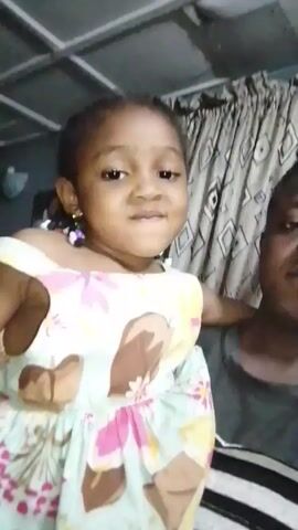 The video shows a young girl with brown skin and short black hair in pigtails. She is wearing a colorful sundress with pink and yellow flowers and brown teddy bear shapes. The girl is smiling and showing her teeth. Behind her is a man with dark skin and short black hair, wearing a gray shirt with white stripes. They are standing in what appears to be a room with light-colored curtains in the background. The room has a metal grid ceiling. The girl is looking directly at the camera and appears to be introducing herself.