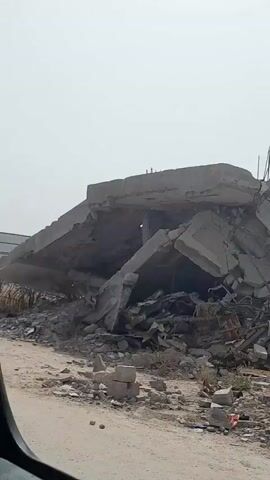 The video shows a scene of destruction. A large pile of rubble dominates the foreground, seemingly the remains of a collapsed building. In the background, partially obscured by the debris, are standing buildings. A man wearing a cap and t-shirt is pulling a cart through the wreckage. The sky is overcast.