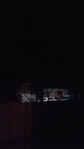 A low-light video shows a dark outdoor scene at night. In the foreground, a reddish-brown wall or fence is visible on the left. Behind it, silhouetted trees and buildings are dimly lit. A small, red light or object is visible in the dark sky above the trees. The video appears to be shaky and has a very low frame rate.