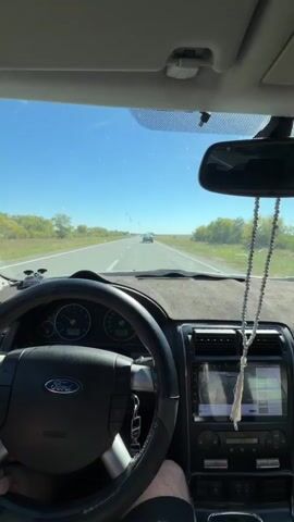 The video shows a first-person perspective from inside a car driving on a sunny day. The driver's hands are not fully visible, but a portion of their arm is. The dashboard and steering wheel of a Ford vehicle are prominent in the foreground, along with a rearview mirror hanging from the windshield with prayer beads. The road ahead is straight and appears to be a highway or main road, with trees and fields on either side. The sky is clear and blue with some minor debris visible on the windshield. Another car is visible in the distance on the road. The car appears to be in motion.