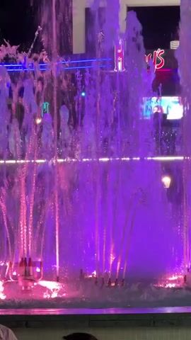A synchronized fountain show features water jets illuminated by pink and purple lights. In the background, a building with signage is visible. A few people are partially visible in the foreground, sitting or standing near the fountain.