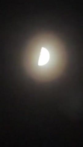 The video shows a half-moon in a dark sky. There is some light around the moon, possibly due to atmospheric conditions. Voices are heard in the background.