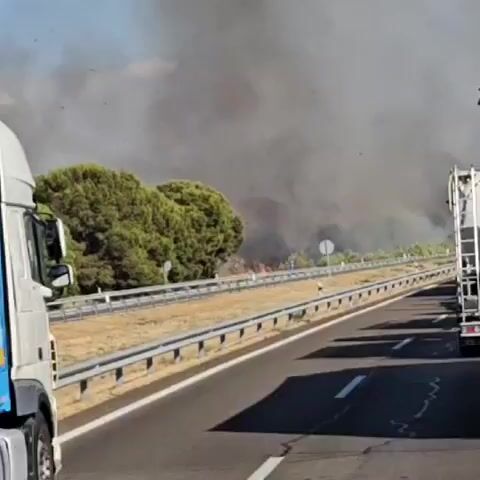 A wide shot of a highway with a plume of black smoke rising in the distance. On the right side of the road, a fire truck is parked. On the left side of the road, a white semi-truck is moving away from the camera. The sky is blue with a few birds flying. The ground is dry and there are trees on the left side of the highway. The smoke is coming from a fire in the distance. The fire appears to be spreading rapidly. The highway appears to be closed due to the fire.