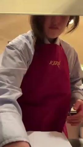 A woman wearing a red apron with the word "Keri" embroidered on it and a light blue button-down shirt rolls up her sleeves in what appears to be a commercial kitchen or bakery. She is standing in front of a counter with a glass display case and several recessed lighting fixtures on the ceiling are visible. The woman has dark hair partially pulled back and is looking directly at the camera with a neutral expression. She methodically adjusts the cuff of her left sleeve, revealing a small tattoo on her wrist. A stack of white papers or notebooks is visible on the counter in front of her. The background includes parts of the kitchen setup, including the metal frames of the glass display case.