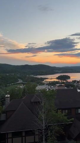 The video shows a view from a high vantage point, looking out over a body of water and a town nestled among rolling hills. The sky is a gradient of blues and oranges, with clouds scattered across the horizon, suggesting either sunrise or sunset. In the foreground, the dark roofs of several houses are visible, partially obscured by trees and foliage. The middle ground reveals a town with buildings clustered together, some appearing to be of traditional architecture. Beyond the town, the tranquil water stretches out, with a small island in the center. Boats are visible anchored near the shore. The overall atmosphere is peaceful and scenic, with the natural landscape dominating the scene. No people or animals are visible in the video.