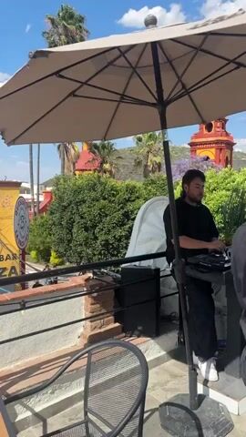 The video shows a view of Peña de Bernal mountain, with the camera panning to focus on the mountain and the sky. The view is from a restaurant or bar terrace with tables and chairs. There is a sign for "Restaurante & Bar Terraza La Vaca Pura" visible in the first frame. The weather is sunny with scattered clouds. There are buildings and trees visible in the midground, in front of the mountain.