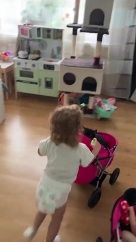 A toddler with curly brown hair, wearing a white t-shirt and a diaper, is pushing a pink toy stroller. A black cat is resting inside the stroller. The child is walking away from the camera, towards a wooden floor. In the background, a multi-level cat tree is visible, along with a toy kitchen and other children's toys. A bright yellow and white shelf unit is on the right side of the frame. The scene appears to be indoors, possibly in a living room or playroom, with natural light coming from a window. The overall mood seems playful and domestic. No text is visible in the media. The location context is Vangaži, Latvia.