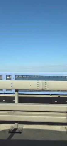A vertical video shows a view from a moving vehicle on a bridge. The foreground consists of highway guardrails made of metal and concrete. Behind the guardrails, there is a blue structure with glass panels, possibly a sound barrier. Beyond the barrier, a body of water with numerous boats docked can be seen. The sky is clear and blue. The video captures the motion of the vehicle as it travels, causing a slight blur in the background.