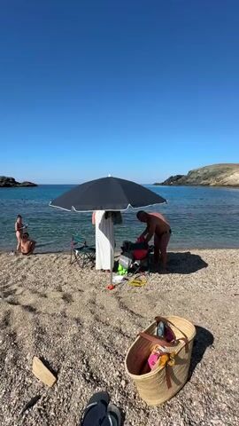 The video shows a sunny day at a pebble beach with clear blue water. Several people are present. In the foreground, a straw bag filled with colorful items sits on the sand, along with a pair of flip-flops. To the left, near the water's edge, two individuals are visible, one crouching and one standing. In the middle ground, a large, dark umbrella provides shade for a setup that includes a cooler and a backpack. A person in a white garment stands near the umbrella, while another person, wearing swim trunks, is interacting with the cooler or backpack. The ocean stretches out to the horizon, with hills visible on the right side of the frame. The sky is a vibrant, cloudless blue.