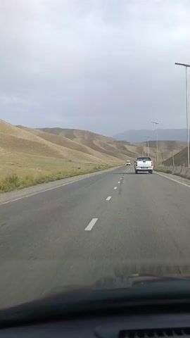 The video shows a view from inside a vehicle driving on a paved road. The road is divided into lanes by white dashed lines. In front of the vehicle, there are two other vehicles: a white pickup truck in the right lane and a white van in the left lane. The landscape on either side of the road consists of dry, rolling hills and sparse vegetation. The sky is overcast with gray clouds. The scene suggests a journey through a somewhat arid, mountainous region under a cloudy sky.