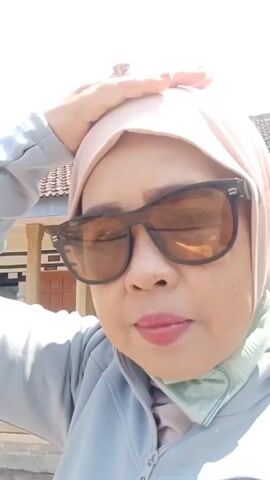 The video features a close-up shot of a woman wearing a light pink hijab and sunglasses. She is also wearing a gray jacket over a light green garment. The woman is outdoors, with a house and a tiled roof visible in the background. The sky is clear and blue, and there are palm trees and other foliage to the left. The woman is touching her head with her right hand. The scene appears to be a sunny day in a rural or suburban area.