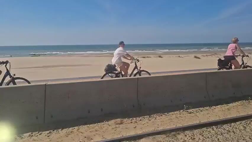 The video shows a view of a beach and the ocean on a sunny day. In the foreground, there is a concrete wall. To the left, a woman on a bicycle rides along a path next to the wall. Further to the right, another person is on a bicycle, and behind them, two people are sitting on a bench. The ocean is visible in the background, with waves gently lapping at the shore. The sand is light brown, and there are some sparse plants growing on the dunes. The sky is clear and blue. The time of day appears to be daytime, likely late morning or early afternoon, given the bright sunlight and the shadows. The overall mood is peaceful and relaxed, suggesting a pleasant day for recreation by the sea. There is no visible text in the media file.