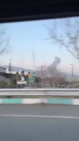 The video shows a view from inside a moving vehicle looking out towards a landscape. The view is partially obscured by the vehicle's window frame at the top. The scene outside includes a highway with a green and white barrier on the side. Beyond the barrier, there's a park-like area with trees, some of which are bare, and grass. In the distance, there are buildings and mountains visible against a light blue sky. At the beginning of the video, smoke is seen rising in the distance.
