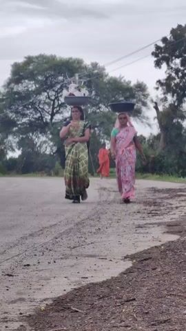 Two women walk along a dirt road. The woman on the left wears a green and white patterned sari and carries a basket of items on her head. The woman on the right wears a pink and white patterned sari and carries a dark-colored basket on her head. A truck with a yellow tarp is visible in the background on the left side of the road. Trees and overcast skies are visible in the background.