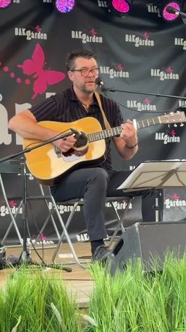 A man sits on a folding chair on a wooden stage playing an acoustic guitar and singing into a microphone. He is wearing glasses, a black and gray striped short-sleeved shirt, and black pants. The background is a black banner with pink butterflies and the text "BAKgarden" repeated. There are lights above the stage. In the foreground, there is green grass.
