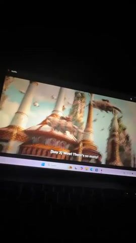 The video displays a laptop screen in a dark room, suggesting it's nighttime or the room is unlit. The screen shows a scene from a movie or TV show, likely "Avatar: The Last Airbender" given the characters and setting.

Initially, the screen shows elaborate, golden-domed buildings with intricate architecture. The first visible subtitle reads "[boy 3] Wow! There's so many!". The scene then shifts to the back of a bald character with a blue and green arrow tattoo on his head, looking towards the buildings. Following this, the camera focuses on an elderly bald man with a white mustache, wearing a red and yellow robe with a beaded necklace, smiling at the camera. The subtitles during this sequence read "[boy 4] Look at all of them!" and the elderly man's line "All of them."

The Netflix logo is visible in the top-left corner of the video player interface. The laptop's Windows taskbar is also visible at the bottom of the screen, showing the "Search" bar and various application icons including File Explorer, Chrome, Microsoft Edge, Spotify, Photoshop, and Netflix.
