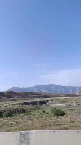 A wide shot captures a rural landscape under a clear blue sky. The foreground is dominated by a concrete barrier, likely the edge of a road or bridge, showing some wear and tear. Beyond the barrier, rolling hills covered in dry, golden grass stretch towards distant mountains. The terrain appears somewhat arid, with evidence of a small ravine or gully cutting through the landscape. The overall impression is one of natural, open space, with a road or man-made structure subtly present.
