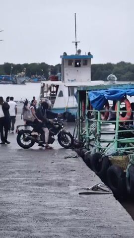 A person is riding on the back of a motorcycle as it is parked next to a ferryboat. The motorcycle has a person driving it and another person on the back, a woman. The motorcycle is facing towards the ferryboat, and the ferryboat is docked at a pier. There are other people visible on the ferryboat and on the pier. The sky is overcast and the ground is wet, suggesting it may have rained recently. The ferryboat appears to be carrying passengers and vehicles. The name "ML EASTER" is visible on the side of the ferryboat, along with a registration number.