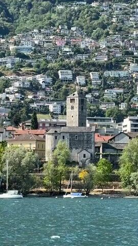 The video displays a panoramic view of a town situated on a steep hillside overlooking a body of water. The town features a prominent stone church with a tall bell tower as its focal point. Numerous buildings, primarily white and modern in design, are clustered on the terraced slopes behind the church, interspersed with lush green trees. In the foreground, the blue water of the lake is visible, with a white sailboat docked near the shore. The overall impression is of a picturesque European lakeside settlement.
