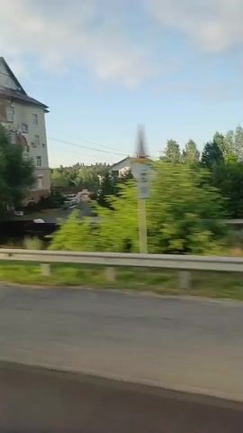The video shows a car driving down a road. On the right side of the road, there are trees and houses. The sky is blue with some clouds. There is a guardrail along the side of the road. The video is taken from inside the car, so the perspective is of someone looking out the window. The scenery is a rural or suburban area. There are no people visible in the video.