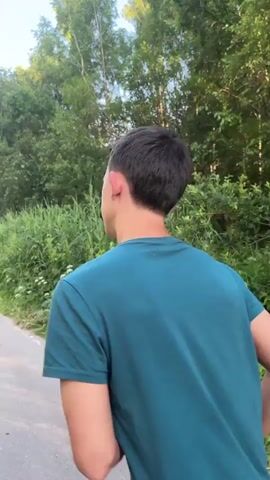 The video shows a person running on a paved road surrounded by trees and vegetation. The person is wearing a teal t-shirt and black shorts and is running away from the camera. The road appears to be in a rural or park-like setting with sunlight filtering through the trees.