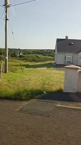 The video shows a roadside view with a dirt road in the foreground. A utility pole with wires is visible on the right side of the screen. Beyond the road, there is tall grass and a field of cut grass in stripes. In the distance, there are houses, trees, and rolling hills under a clear sky. A white van with its doors open is parked on the side of the road near a fence. A white house with a stone facade and a chimney is also visible among the greenery.
