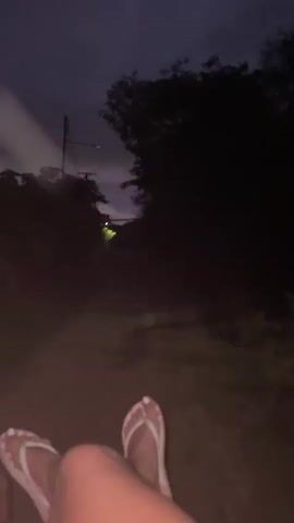 The video begins with a close-up of a person's feet wearing pink flip-flops, resting on a reddish-brown dirt path. The background shows a dark, overcast sky and silhouetted trees, suggesting it is either dusk or night. A dirt path leads into the darkness, with some faint green lights visible in the distance. As the video progresses, the scene becomes darker, and the faint lights remain visible. The camera appears to be stationary, looking forward along the path.