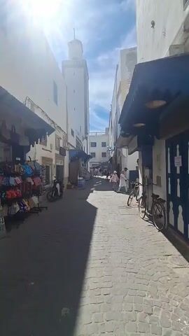 The video shows a narrow cobblestone street lined with buildings. On the left, a stall displays colorful bags. A motorcycle is parked in front of the stall. In the distance, a white minaret rises above the buildings. Several people are walking and sitting along the street. A bicycle is parked on the right side of the street. The sun is shining brightly, casting shadows on the street. The sky is blue with some clouds.