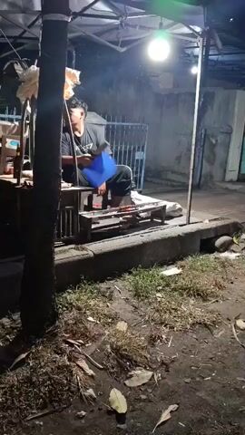 The video shows a young person sitting at a food cart at night. They are wearing a black t-shirt and dark pants. The food cart is lit by a bright overhead light, and there are some items on display, including skewers of food. The person appears to be looking to their right. The surrounding environment is dark and appears to be a street or alleyway, with some vegetation and debris on the ground.