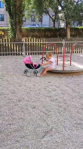 A young girl with blonde hair, wearing a pink shirt and pink shorts, is sitting on a merry-go-round in a playground. She is holding the handle of a pink doll stroller and appears to be pushing it. The merry-go-round is red and is surrounded by light-colored gravel. A wooden fence is visible in the background, along with trees and a building. The overall scene suggests a playful and innocent moment.