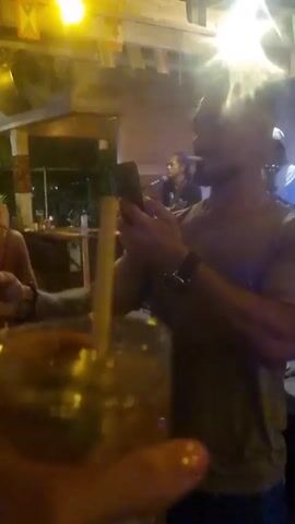 The video shows a man in a beige shirt and dark pants taking a selfie with a phone at an outdoor venue with live music. He's holding a drink in his left hand. A band is visible on a stage in the background. The scene is dimly lit, with bright overhead lights causing glare. Other people are present, some dancing near the band, others seated at tables. The music appears to be reggae.