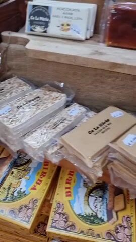 The video shows a display of food items for sale, likely at a market. The items include various types of nougat (torrons), chocolate bars, and jars of preserves or honey. The nougat bars are packaged individually in clear plastic. Some of the nougat appears to have nuts and dried fruit in it. There are also several bars of what looks like solidified honey or fruit paste. In the background, there are jars with dark contents, possibly jams or honey. The overall impression is of a food stall offering artisanal products.