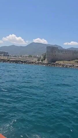 A boat is moving across the water. The camera is pointing towards land. On the land is a large stone structure that appears to be a castle or fortress. Behind the castle, there are buildings and mountains. The sky is blue with some white clouds. The water is a deep blue-green and has some ripples. The front edge of the boat and a person's hair are visible at the bottom of the frame.