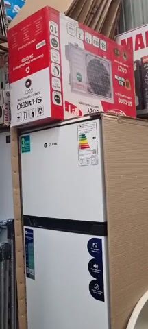The video pans across a market stall filled with various household appliances and goods. In the foreground, a white refrigerator with an energy efficiency label is prominently displayed, stacked behind a large cardboard box. On top of the refrigerator box, a boxed air conditioner unit is visible. To the right, a shelf displays a boxed electric kettle. Above the merchandise, a sign with Cyrillic text and the number "12-53" is attached to the wall. Further to the right, several rolled-up carpets and other items are stacked, with two men standing in the background, one wearing a blue shirt and the other a dark shirt. The market appears to be an indoor or covered space, with a ceiling visible at the top of the frame.