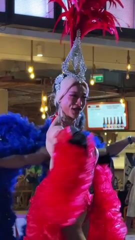 This video captures a vibrant drag performance in Baan Chaweng, Thailand. The central subject is a drag queen dressed in a dazzling red sequined costume, complete with a dramatic headdress adorned with red feathers and a silver crown. Her outfit features a plunging neckline, a black corset, and a voluminous red feather skirt that flares out around her. She wears black gloves and a choker necklace.

To her right, another performer in a blue sequined outfit with blue feather accents is visible. Both performers appear to be engaging with the audience, with smiles and expressive movements. The drag queen in red is particularly animated, with her mouth open in a song or exclamation, and her arms extended.

The setting appears to be an indoor venue, possibly a bar or entertainment space, with ambient lighting from decorative lamps overhead. In the background, patrons can be seen seated at tables, suggesting a live audience for the performance. The lighting suggests it is evening.

A rainbow-colored banner is partially visible in the bottom left corner, hinting at a Pride or LGBTQ+-friendly event. The overall atmosphere is celebratory and energetic, as is typical of a drag show. The visible text on the badge worn by the main performer is a circular shape with a "7" inside it.