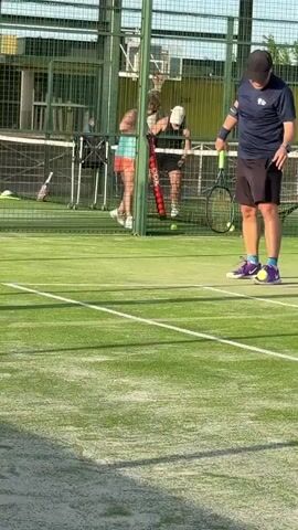 Two people are visible on a green padel court. The person on the right is wearing a dark blue shirt and black shorts, holding a padel racket with a bright green handle and a yellow ball. The person on the left is wearing a turquoise tank top and an orange skirt, also holding a padel racket. They are standing near a green mesh fence. In the background, several other people are visible behind the fence, some of whom appear to be watching the game. The court has white lines marking its boundaries. The lighting suggests it is daytime, likely outdoors. The focus is on the two players in the foreground, with the person on the right in sharper focus.