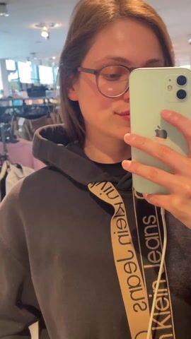 A young woman is holding up a light green Apple iPhone and taking a selfie. She’s wearing round eyeglasses and a black Calvin Klein Jeans hoodie. She puckers her lips slightly in a kissing expression. The background suggests she is in a retail store. The lighting is bright, with numerous overhead lights visible. A white cable is connected to the phone.